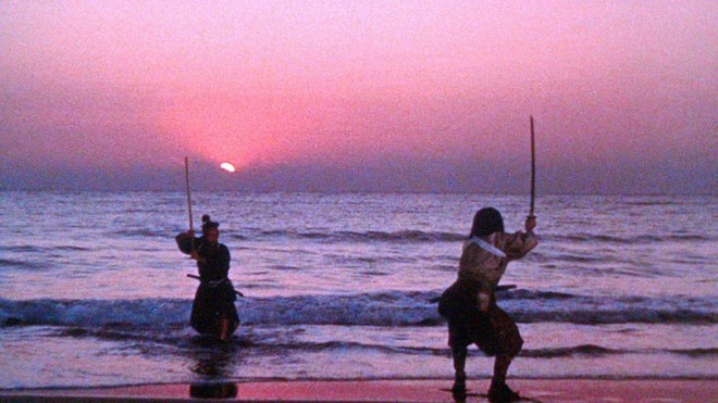 Two samurai face off with raised swords at the shoreline with the sun low on the horizon